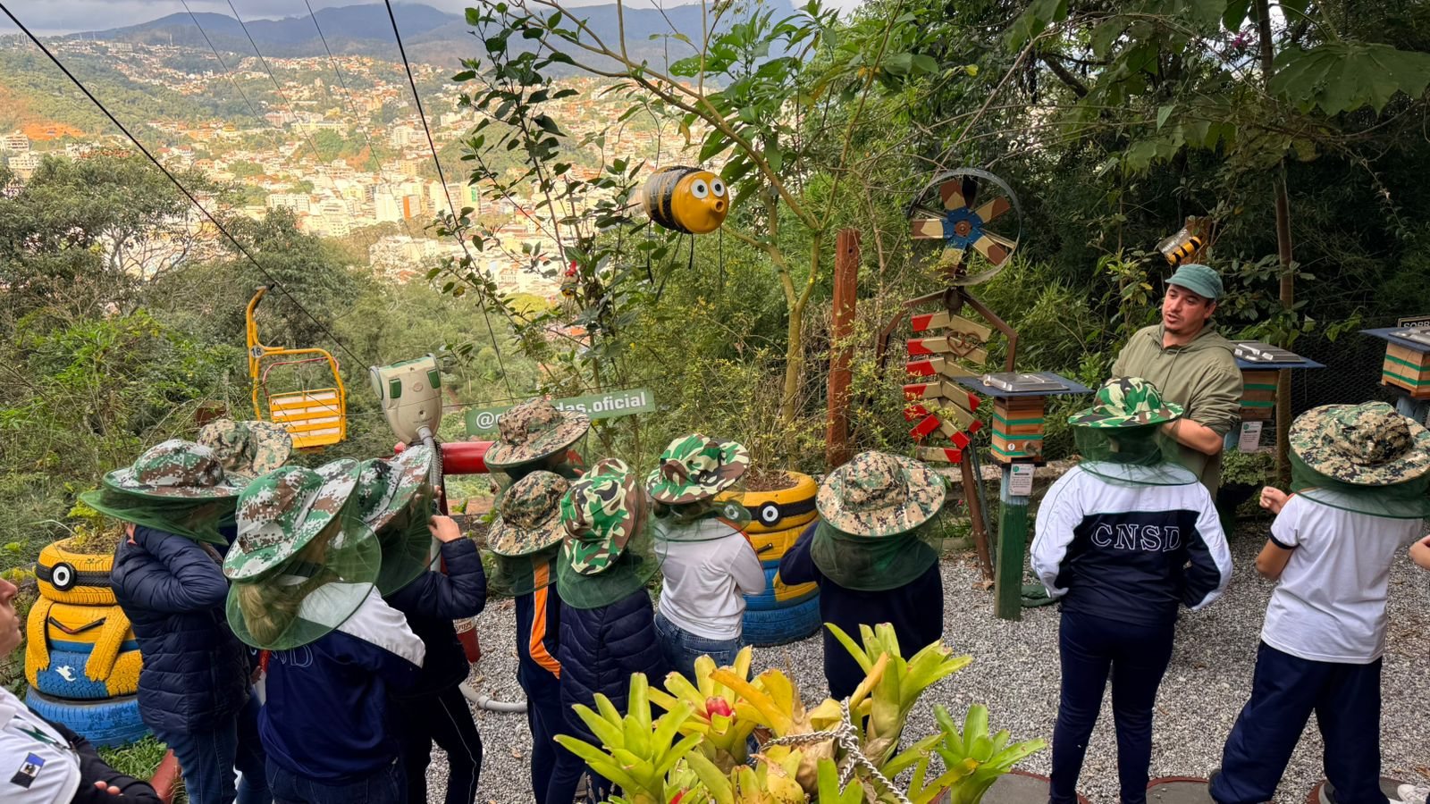 Estudantes de chapéu de apicultor no meliponário da EcoModas, com a cidade ao fundo