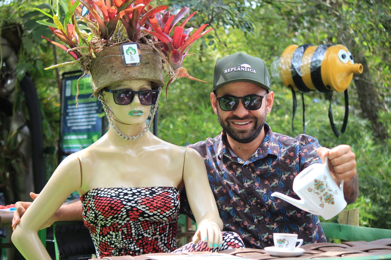 Visitante servindo chá ao lado de um manequim decorado com plantas no jardim da EcoModas
